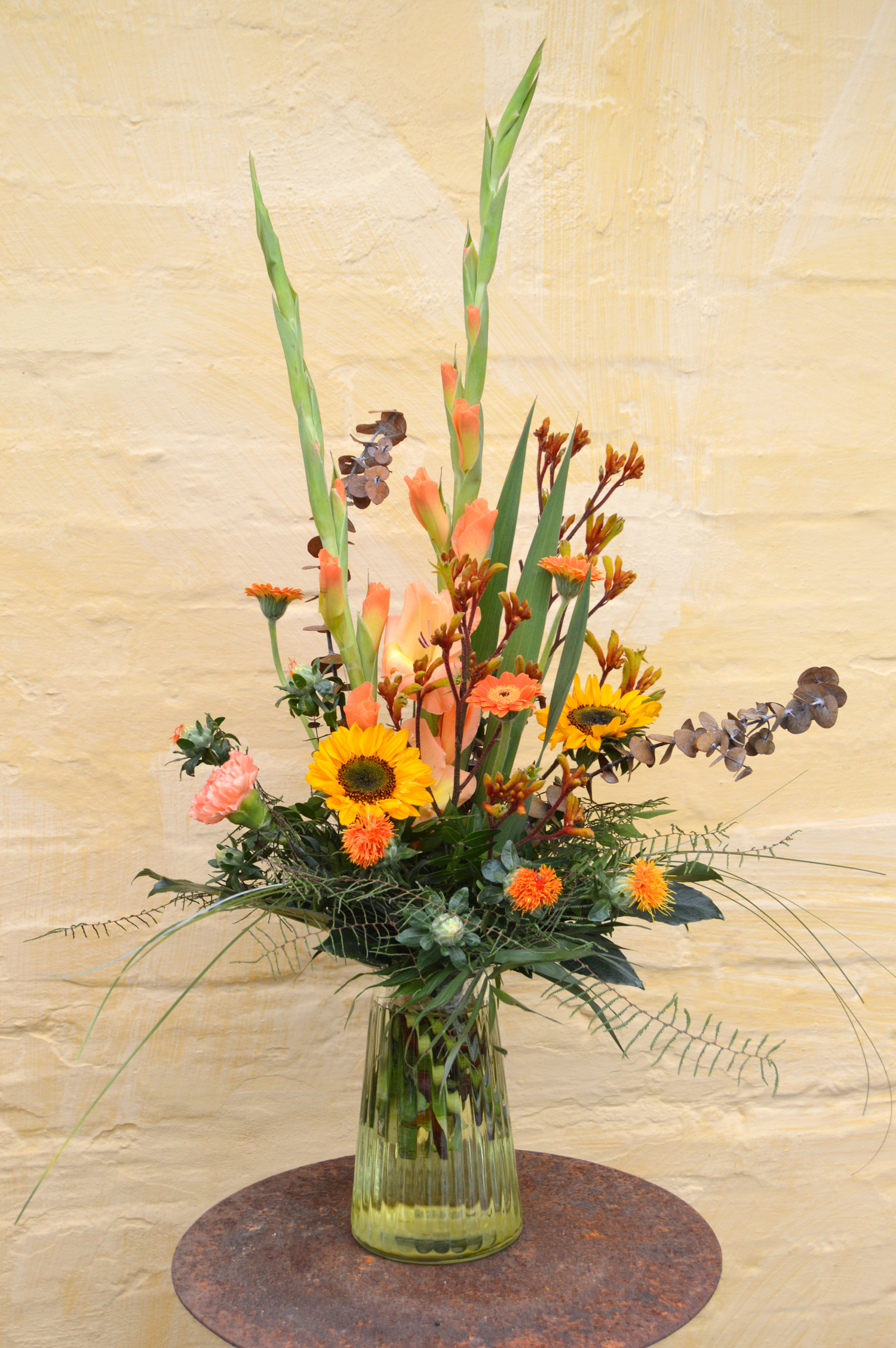 Høj buket med blomster i varme og gyldne nuancer, som orange, gul og rød. Buketten er bundet med et livligt og vildt udtryk. 