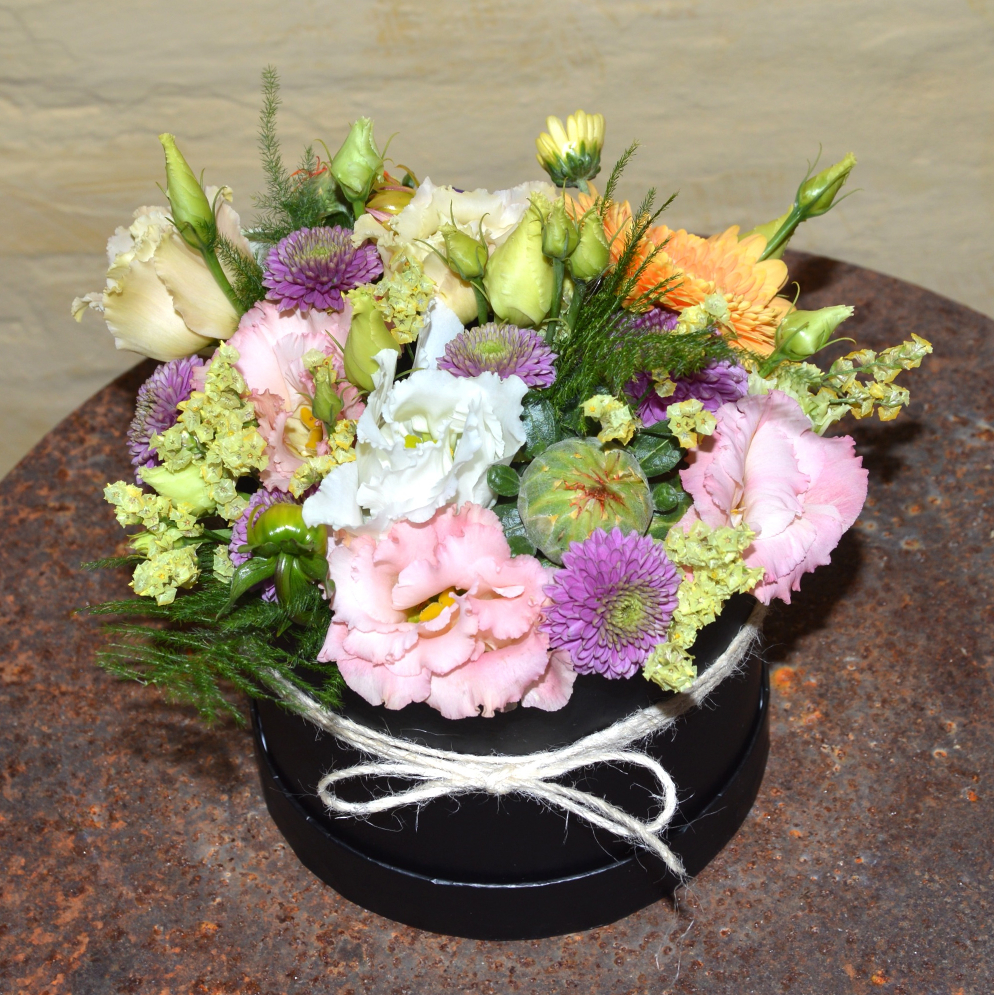 Blomsteriets Hatteæske med rosa nelliker, lilla asters, roser og grønt i sort rund æske.