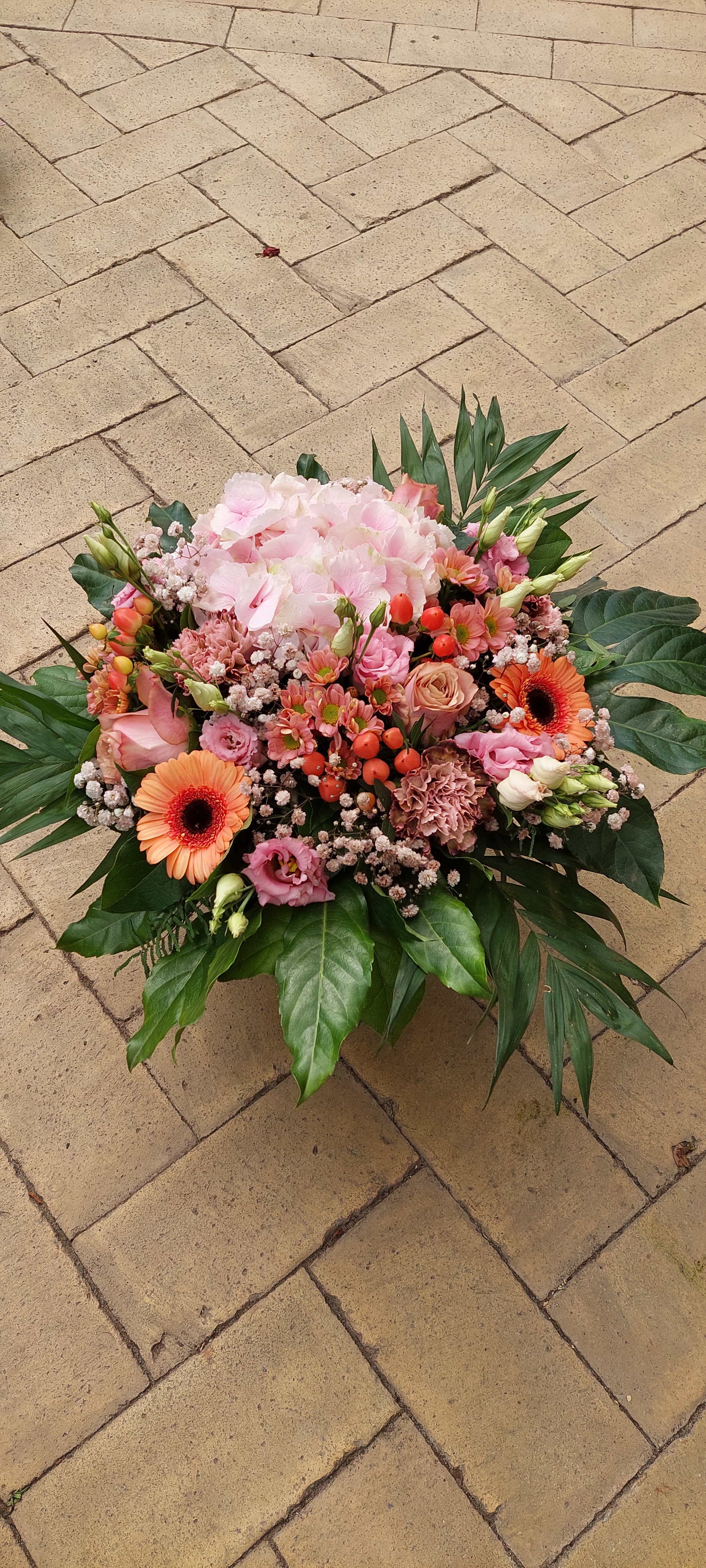 Tæt og rund buket med blomster i forskellige størrelser og former. Den er bundet i pink, røde og lyse nuancer.