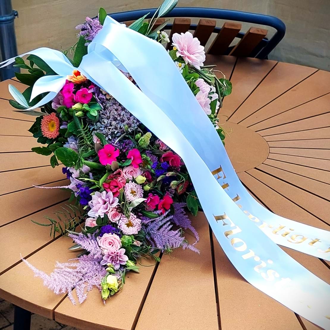 Bårebuket med et vildt og naturligt udtryk. Den er bundet med blomster i gule, blå og lilla nuancer.