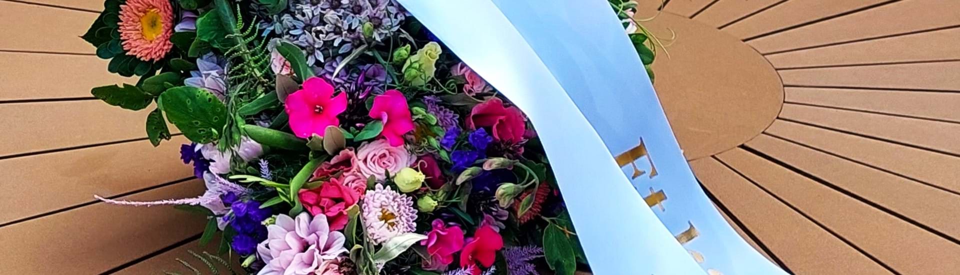 Bårebuket med hvidt bånd er bundet med blomster i lilla, pink, lyserøde og blå sensommerfarver.